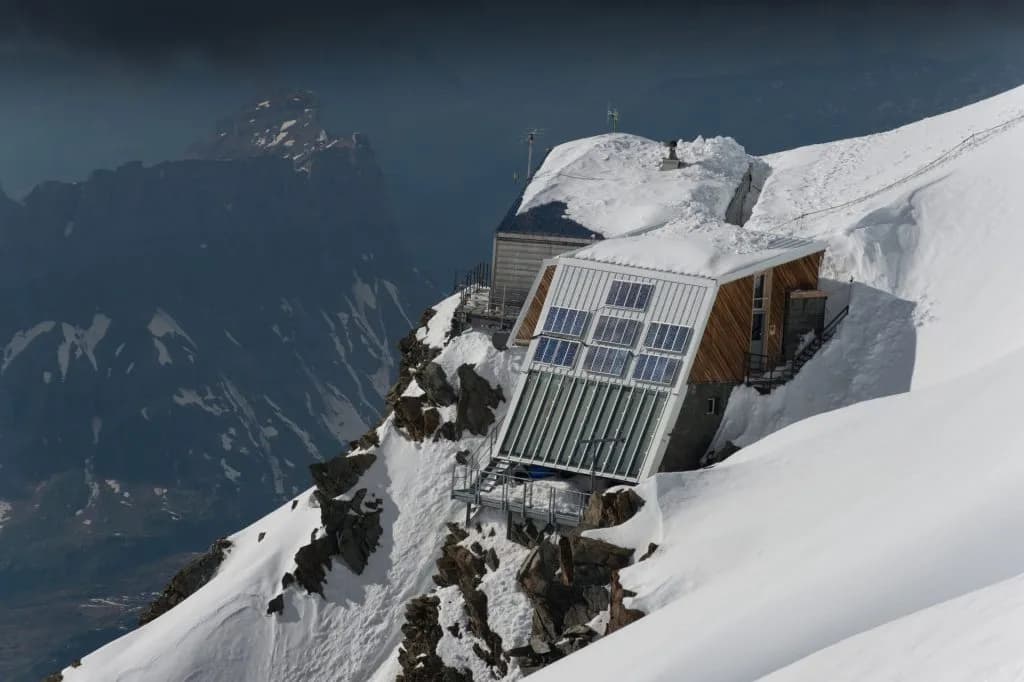 Mountain hut with solar panels clinging to a steep, snow-covered slope overlooking dark peaks.