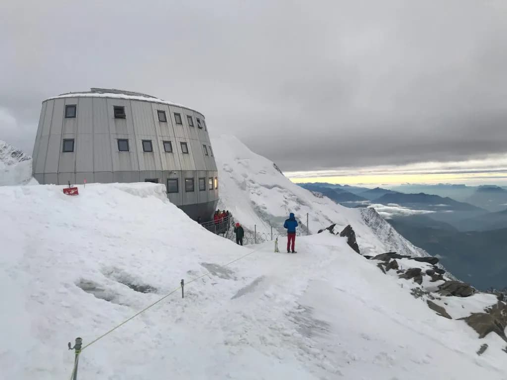 Modern circular mountain hut on snowy peak overlooking layered mountain ranges under cloudy sky