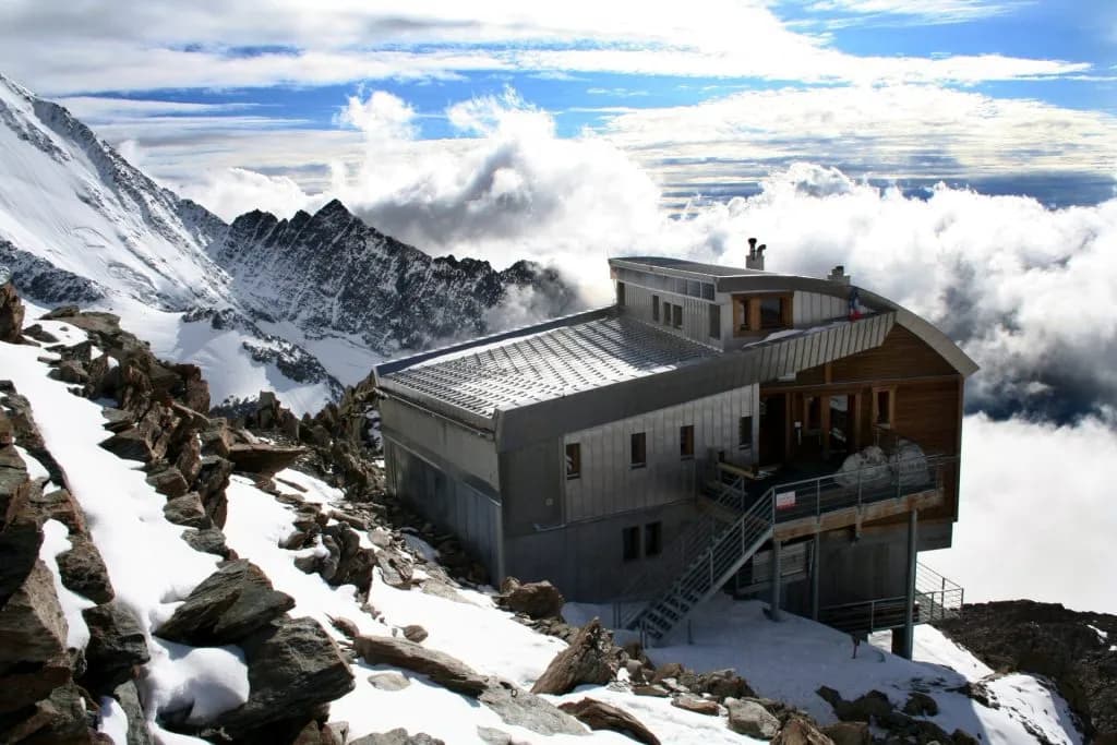 Tête Rousse Mountain Hut in snow surrounded by rocky terrain and high alpine peaks above the clouds.