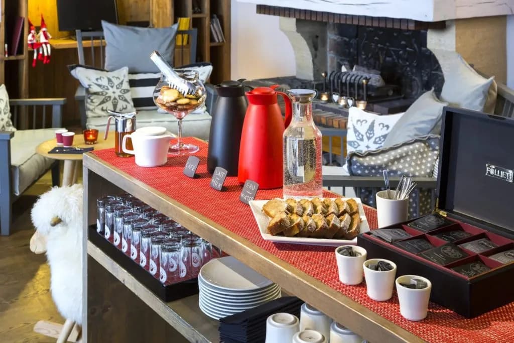 Self-serve beverage and snack station with coffee, milk, water, cookies, and cake near a stone fireplace.