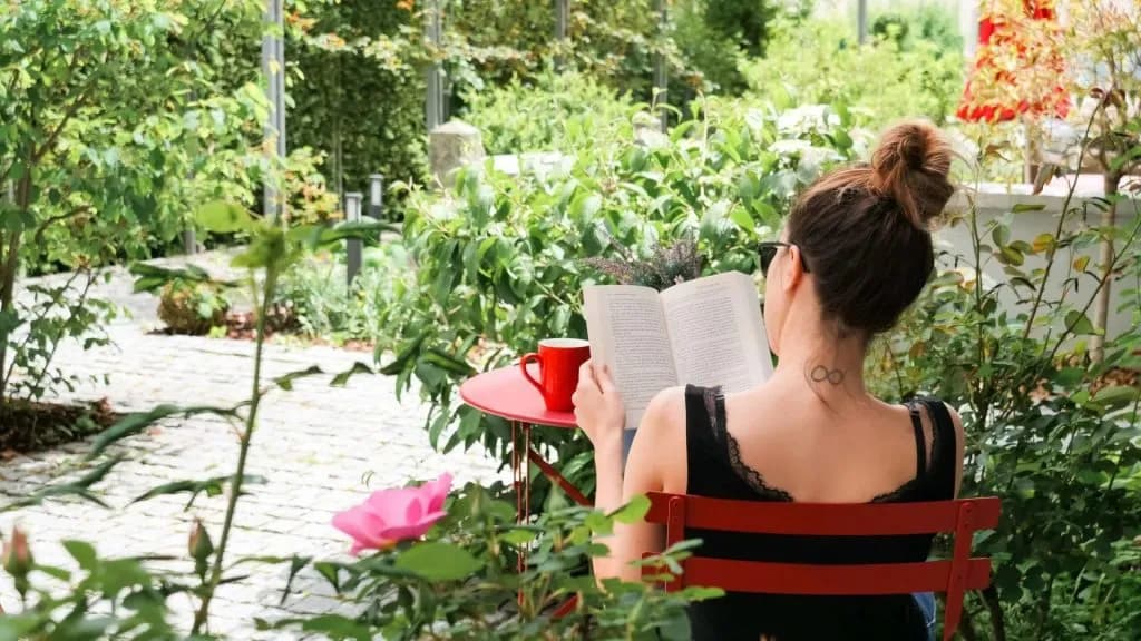 Woman reading a book with a red mug in a lush green courtyard with pink roses.