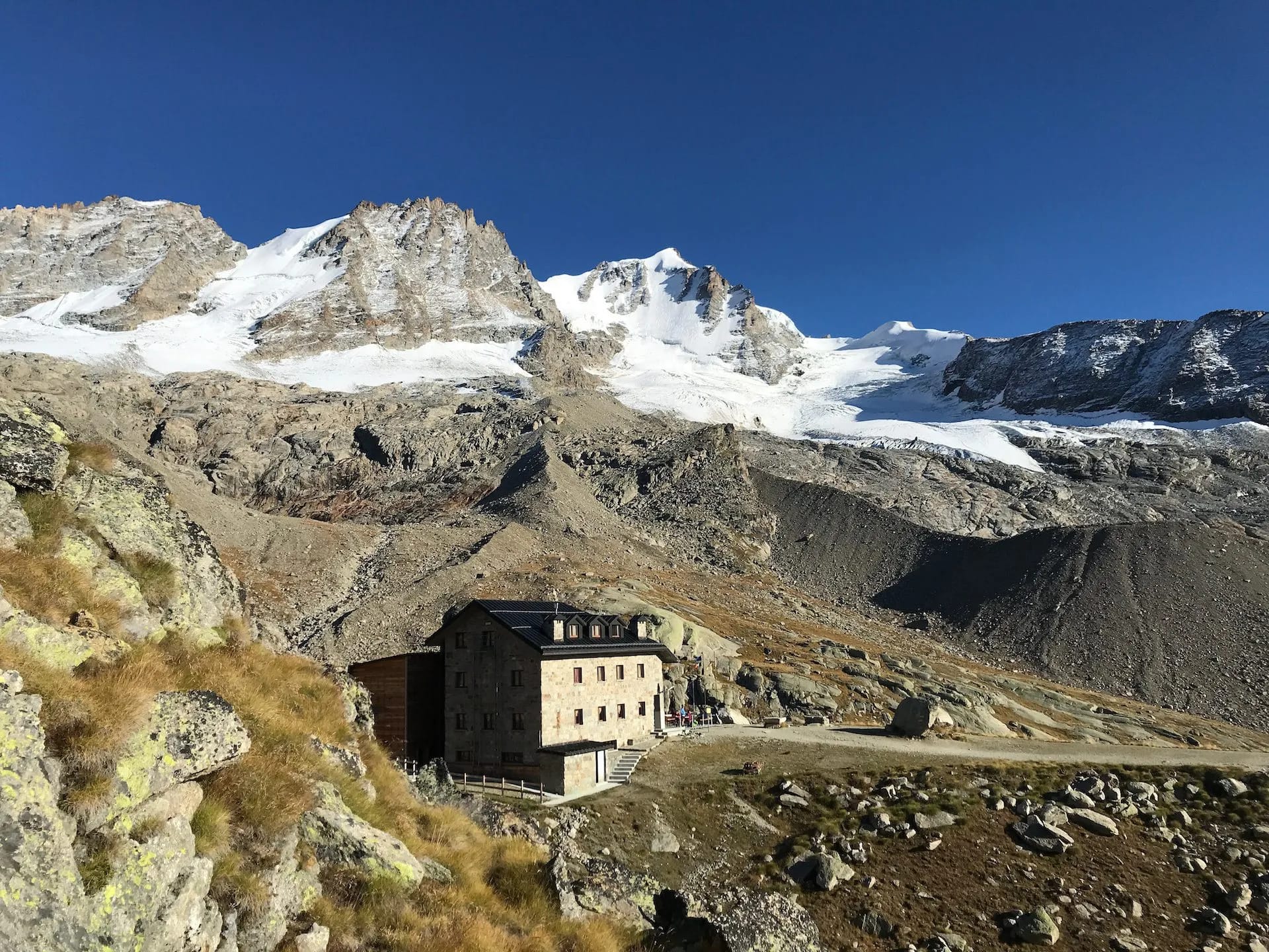 Rifugio Chabod stone mountain hut below snow-capped peaks under clear blue sky.