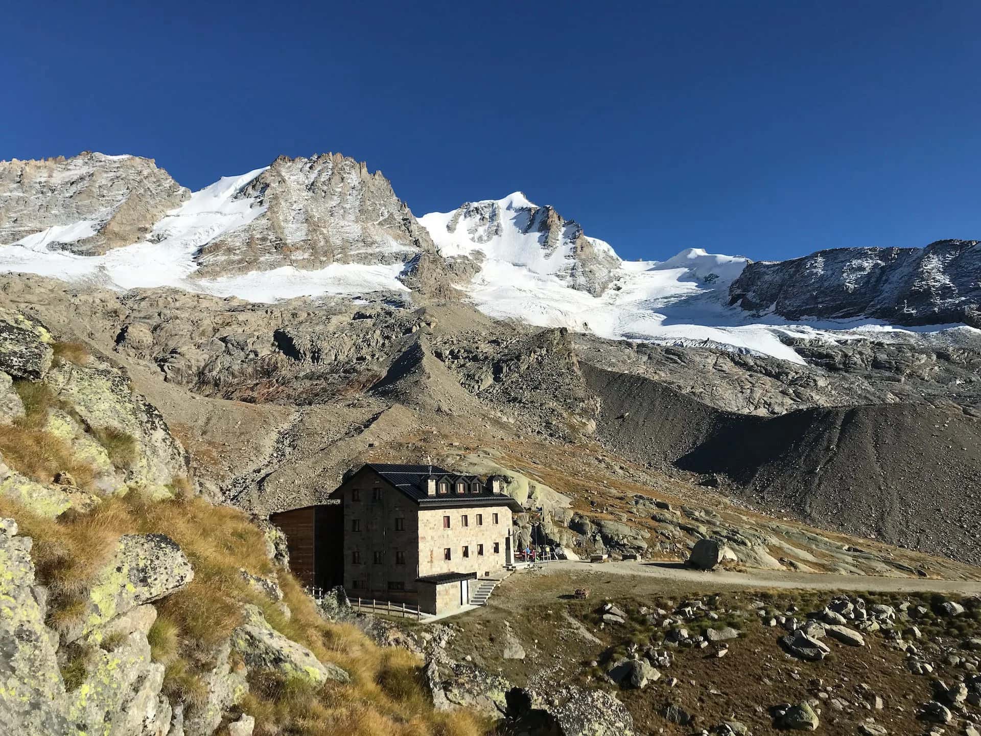 Rifugio Chabod stone mountain hut below snow-capped peaks under clear blue sky.
