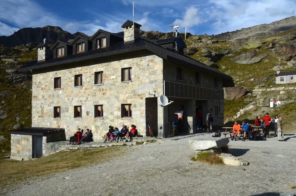 Rifugio Chabod stone mountain hut with hikers resting outside against rocky alpine backdrop.